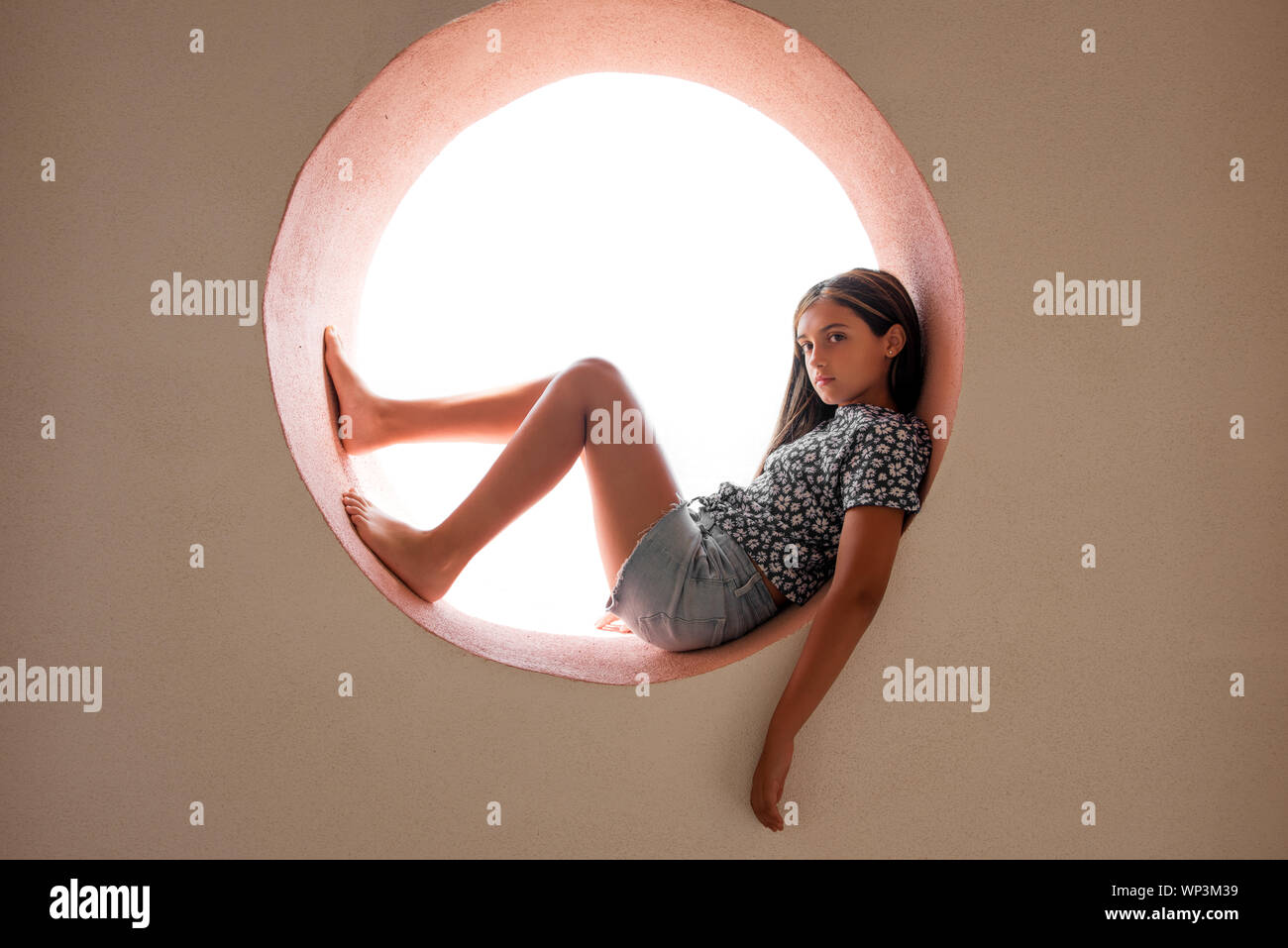 Young barefoot pretty little girl resting in a circular opening in a cement wall backlit by bright light looking thoughtfully to the side of the camer Stock Photo
