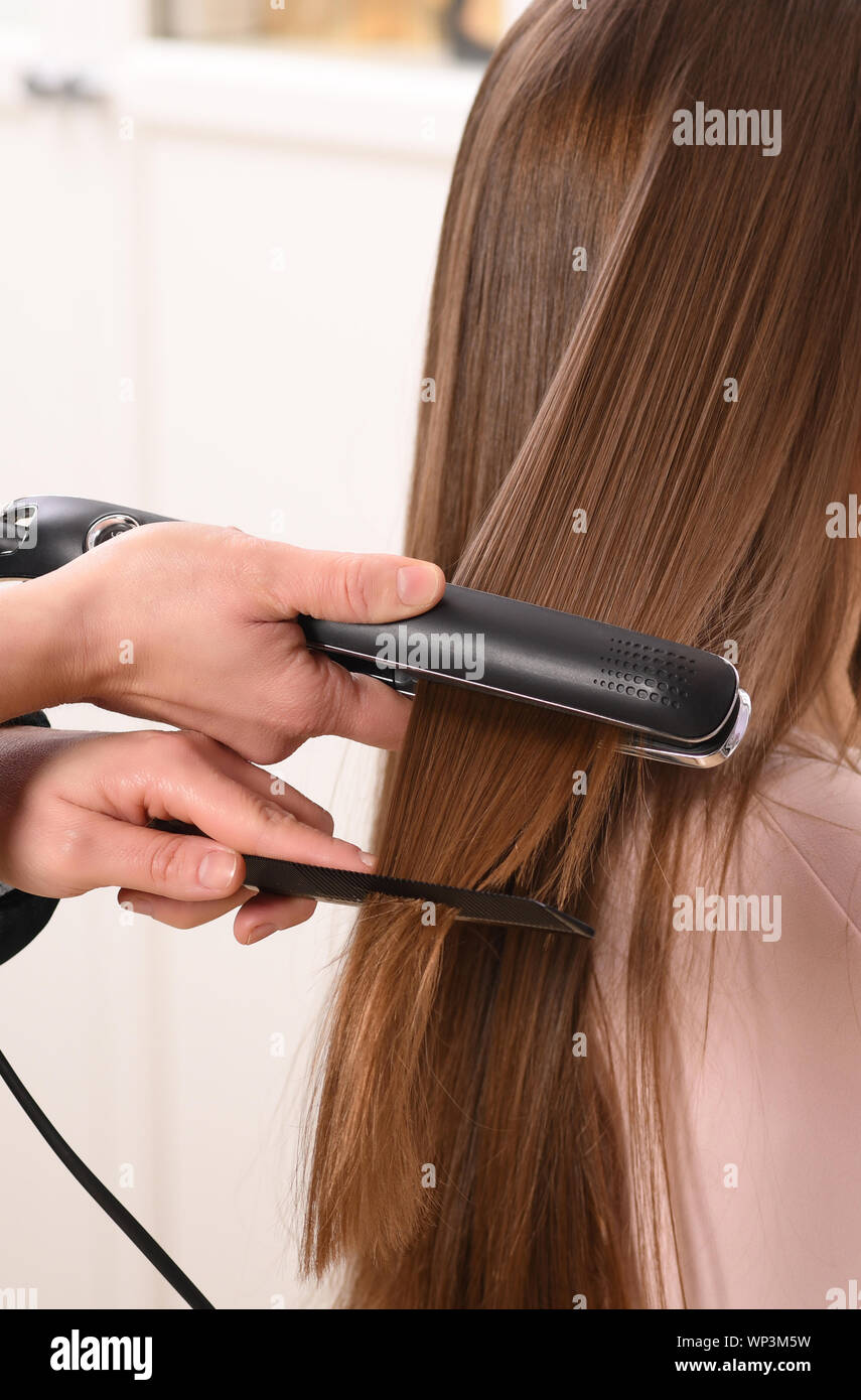 Close up on unidentifiable persons hands using black hair straightener and comb on womans brown hair Stock Photo