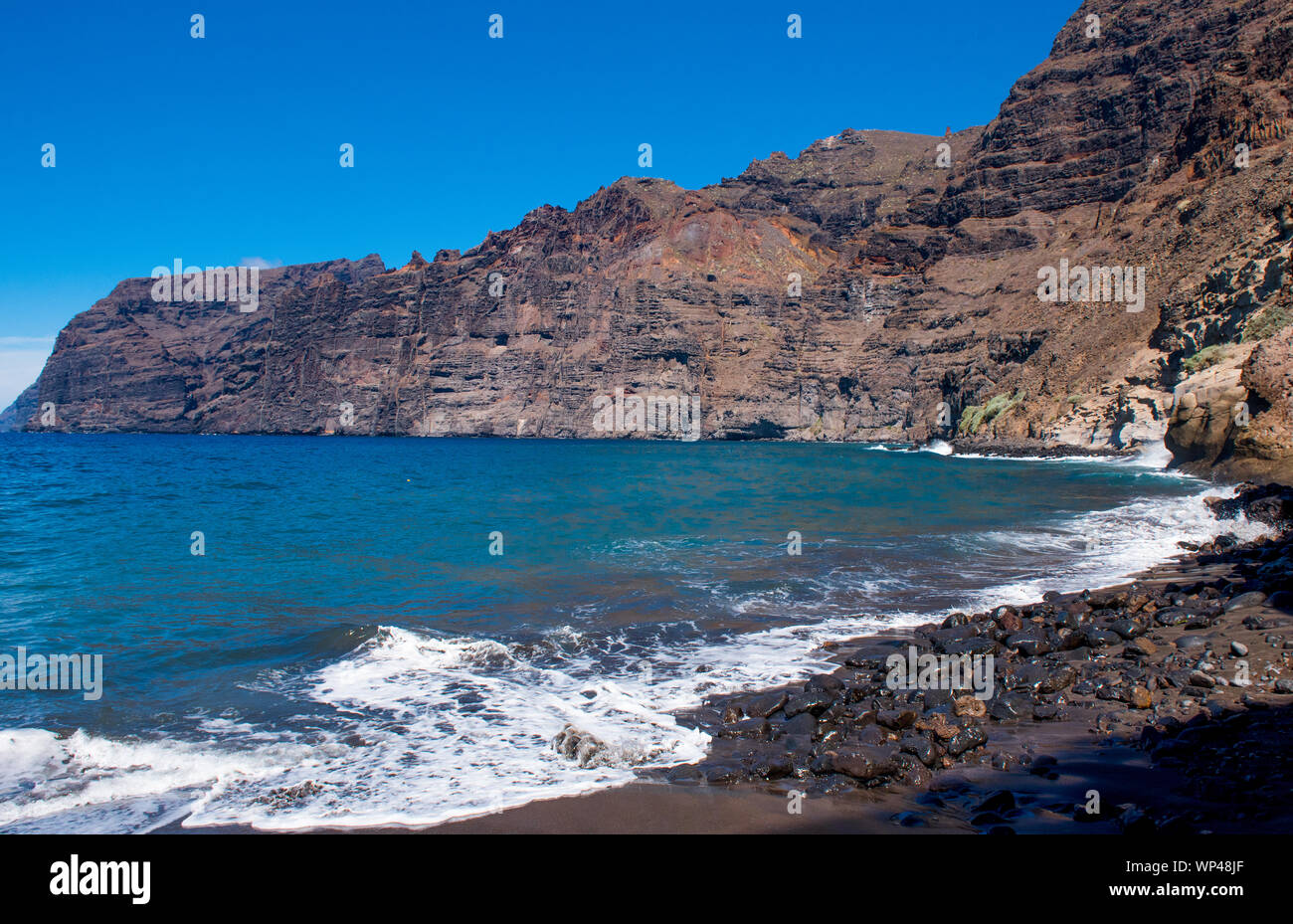 The dramatic towering volcanic cliffs of Los Gigantes, Tenerife, Canary Islands.  Black stony sand beach, breaking waves blue Atlantic Ocean and sky. Stock Photo