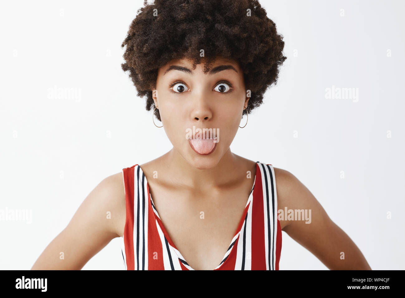 Close-up shot of funny and playful immature stylish woman sticking out tongue and popping eyes at camera fooling around and showing temper while stand Stock Photo