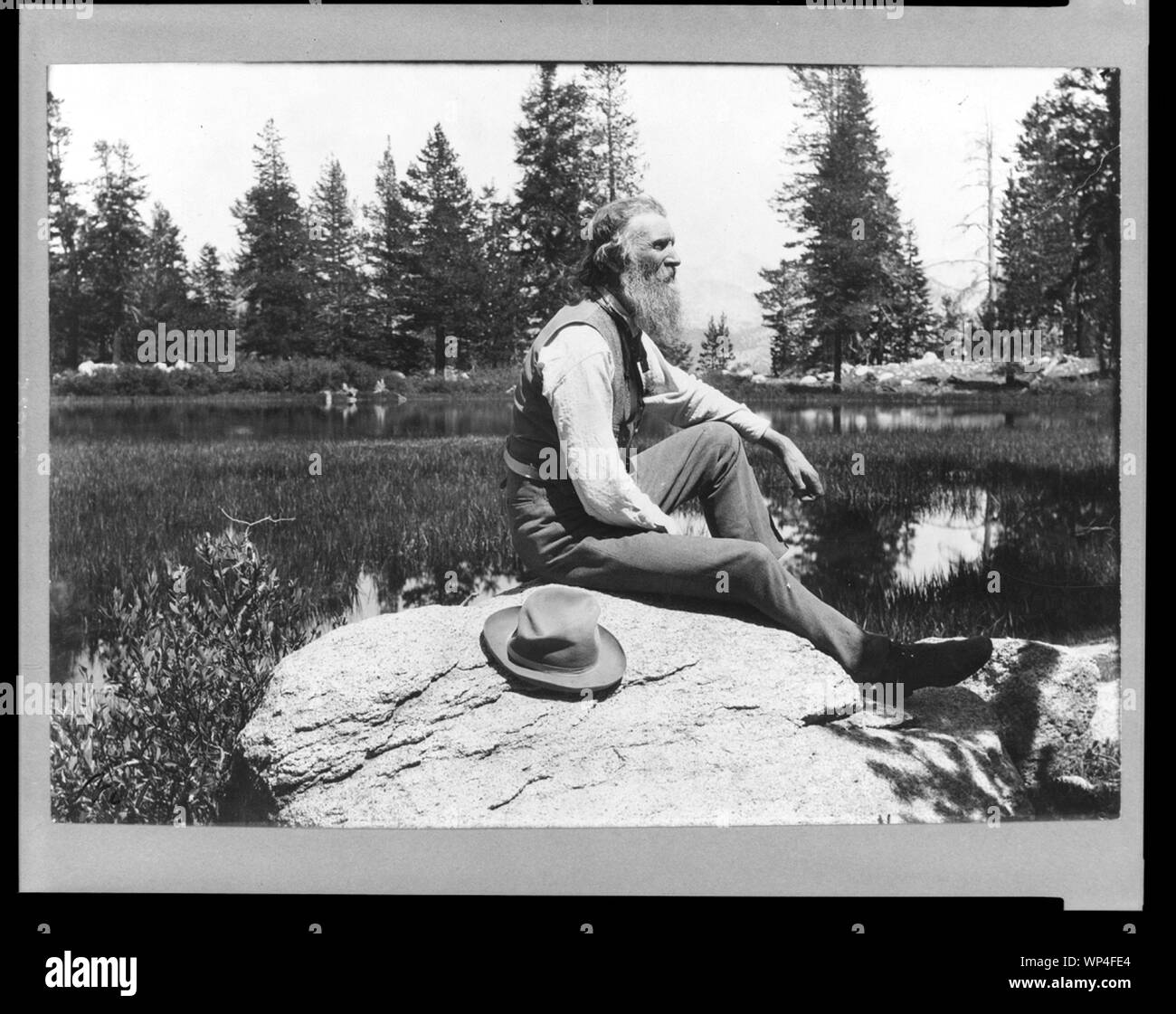 John Muir, full-length portrait, facing right, seated on rock with lake and trees in background Stock Photo