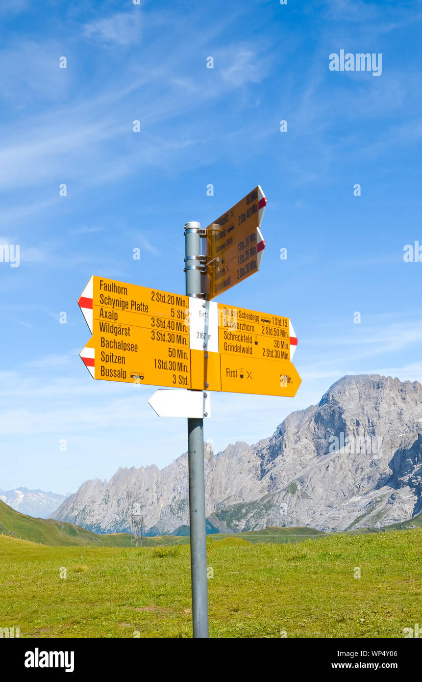 Yellow information sign in First, Switzerland giving distances and directions for hiking in the Swiss Alps. Popular paths by Grindelwald leading to Bachalpsee. Summer Alpine landscape in background. Stock Photo