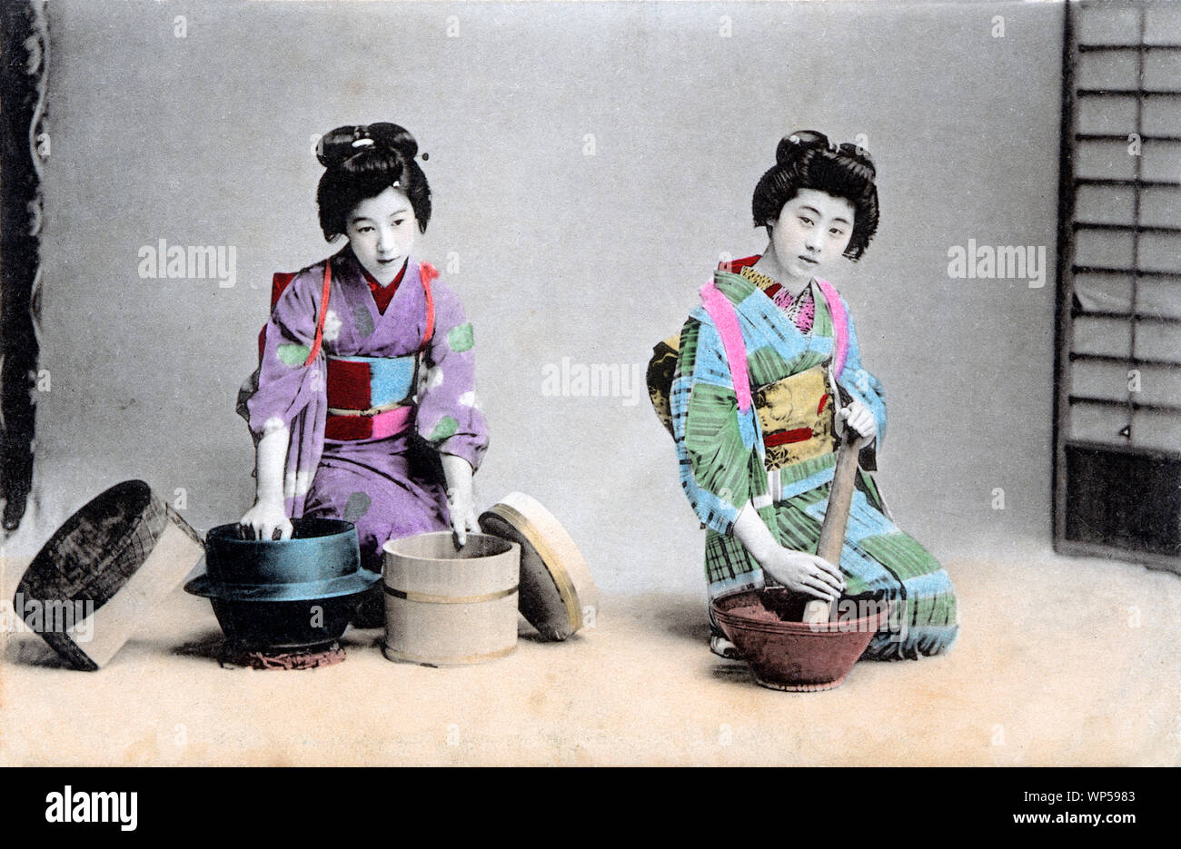 [ 1910s Japan - Japanese Kitchen Work ] —   In this studio photo of daily kitchen work, two women in kimono show typical Japanese kicthen dusties.  The woman on the right is grinding something in an earthenware suribachi (mortar). The other is holding a tetsunabe (iron pot) for cooking rice and an oke (wooden bucket).  20th century vintage postcard. Stock Photo