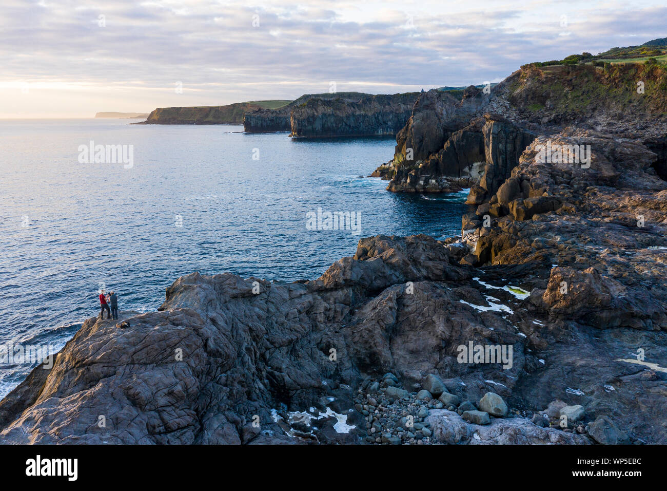 Two photographers take pictures of the sunrise at the rocky coastline of Ponta do Mistério at Terceira Island in the Azores. The morning light hits gi Stock Photo
