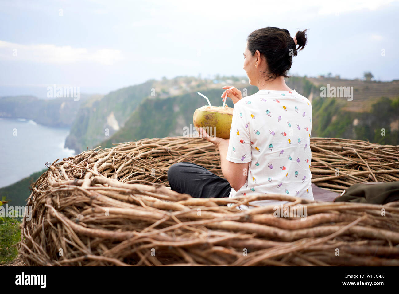 Young Woman Taking A Break And Drinking A Coconut Inside A Nest In Front Of The Sea, Bali, Indonesia Stock Photo