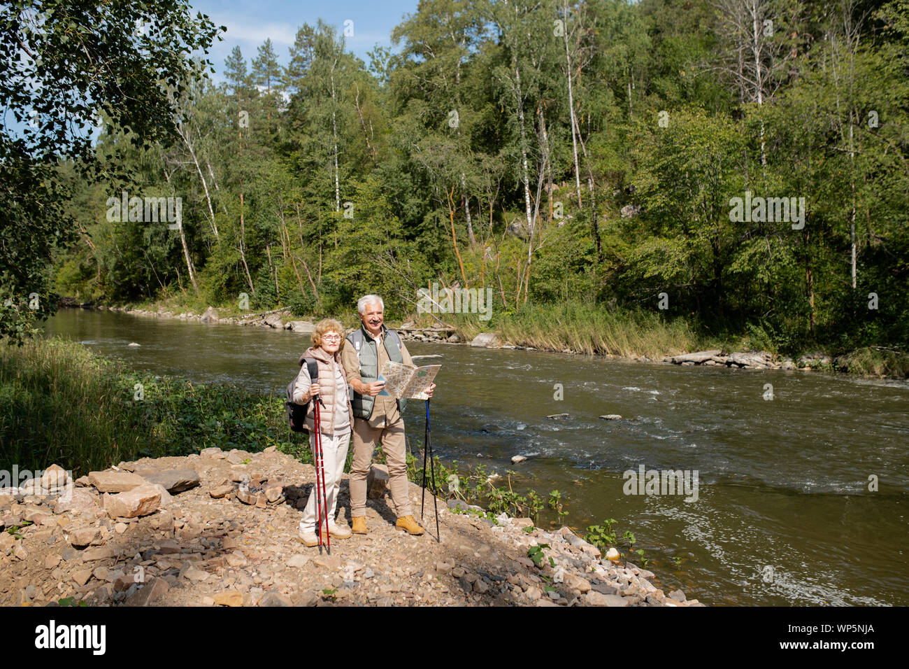 Senior couple with map standing by forest river while going to find way back Stock Photo