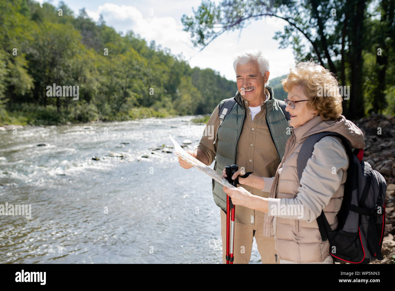 Active senior couple with backpacks looking at map while standing by large river Stock Photo