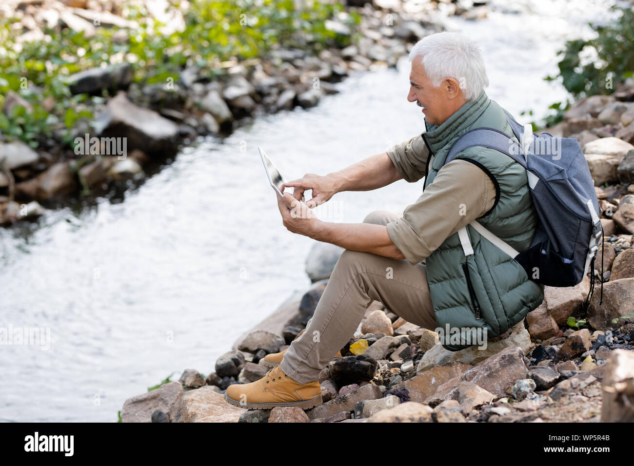 Aged man in activewear pointing at touchpad display while sitting on river bank Stock Photo