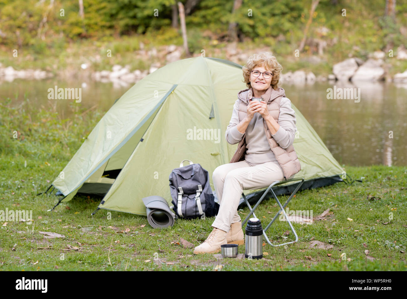 Mature smiling woman holding hot drink while sitting on small tourist chair Stock Photo