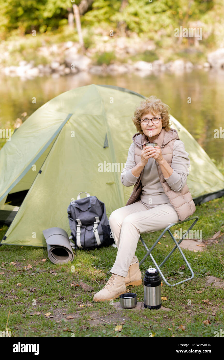 Active mature woman sitting on small chair and having tea in natural environment Stock Photo
