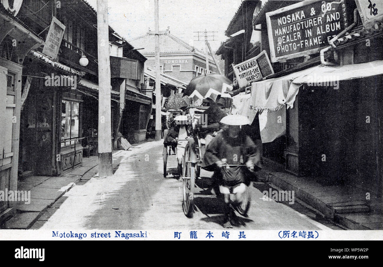 [ 1900s Japan - Rickshaws in a Nagasaki Shopping Street ] —   Two rickshaw pullers pass in front of the album and photo frame shop of Nosaka & Co. on Motokagocho, Nagasaki City.  20th century vintage postcard. Stock Photo