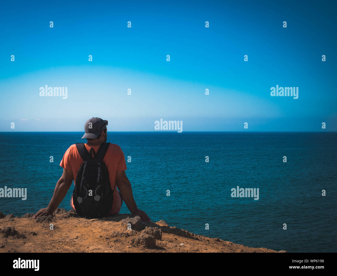 man with cap and backpack sitting on a cliff watching the sea Stock Photo