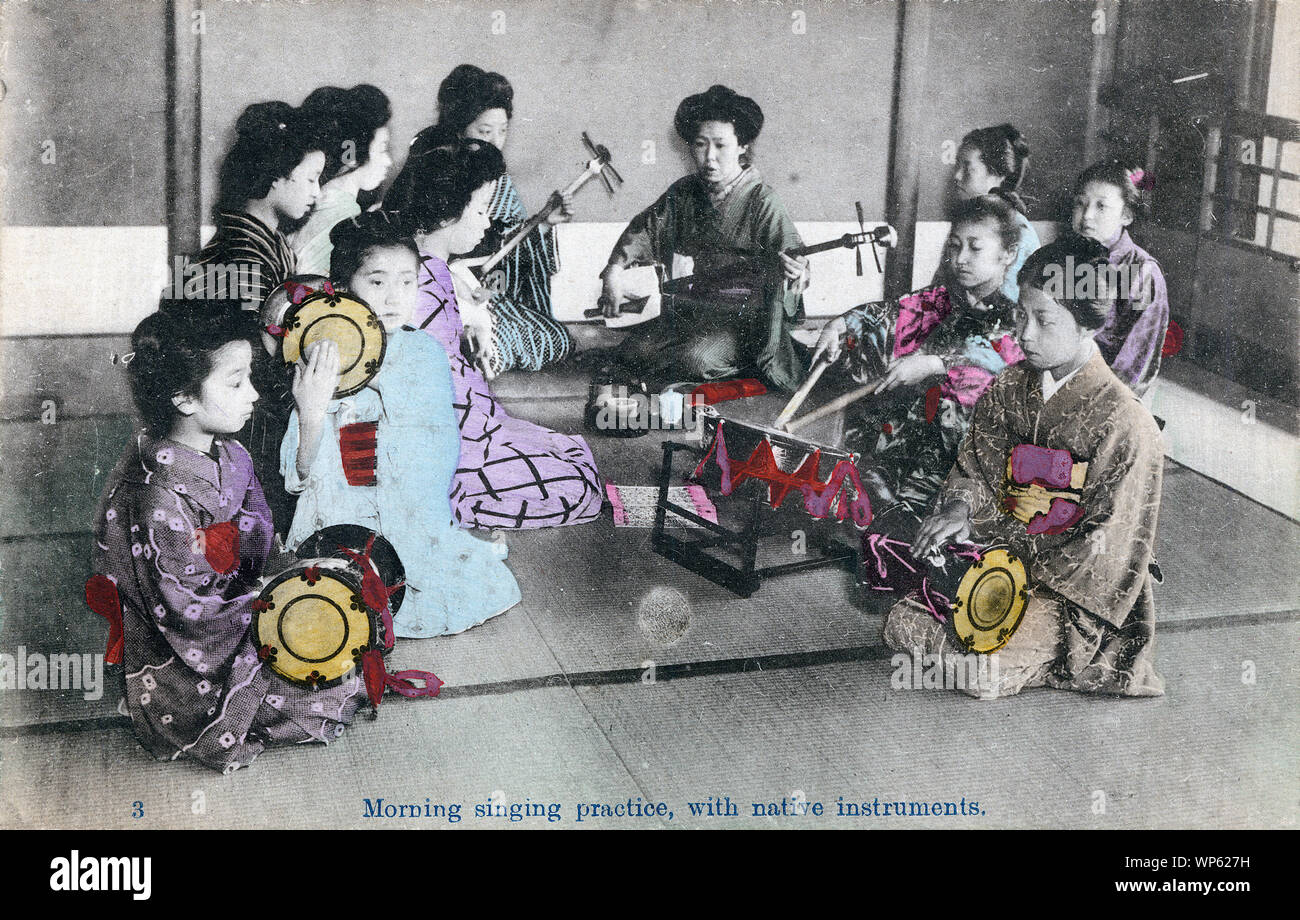 [ 1900s Japan - Traditional Japanese Music Lesson ] —   Girls in kimono and traditional hairstyles are practicing singing and playing Japanese musical instruments  A tsutsumi (also tsuzumi) shoulder drums, shamisen, a three-stringed musical instrument, and a taiko drum known as ootsutsumi can be seen. The teacher is sitting in the back.  20th century vintage postcard. Stock Photo