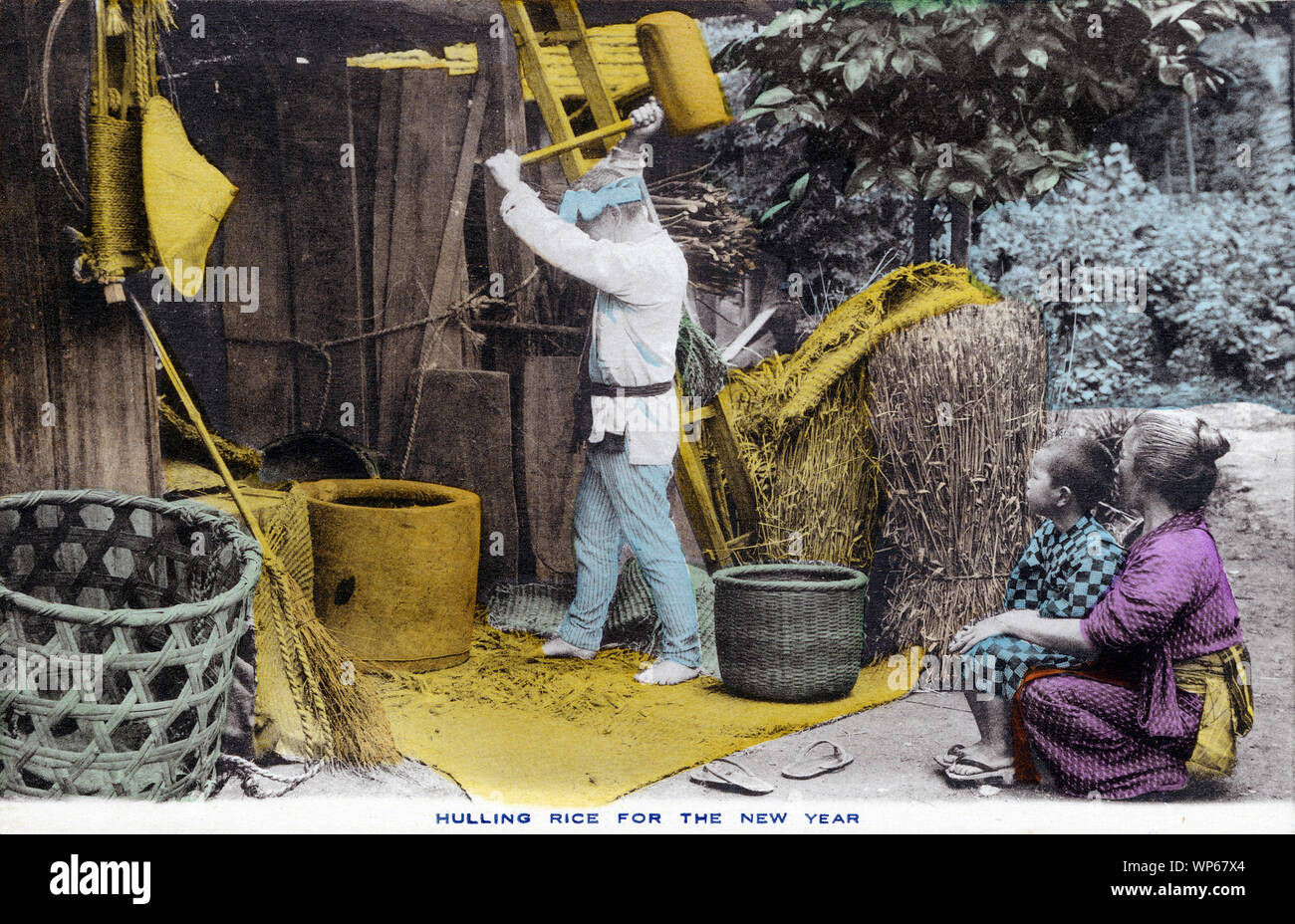 [ 1920s Japan - Japanese Farmer Hulling Rice ] —   A farmer is hulling rice while his wife and son, both wearing a yukata, look on.  This postcard is from a series about Japanese farming, called Farmer Life in Japan.  20th century vintage postcard. Stock Photo