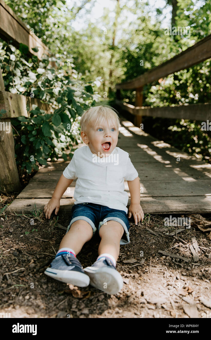 Cute Little Blond Haired Toddler Boy Kid Child Sitting and Laughing in Front of Wooden Bridge Over a Creek at the Park in the Forest During Summer Stock Photo