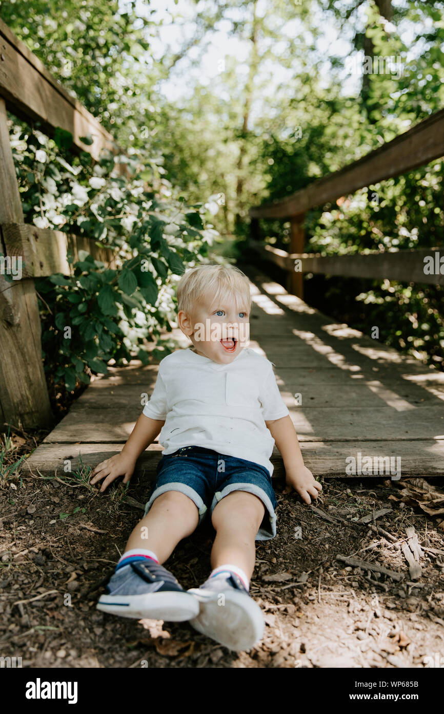 Cute Little Blond Haired Toddler Boy Kid Child Sitting and Laughing in Front of Wooden Bridge Over a Creek at the Park in the Forest During Summer Stock Photo