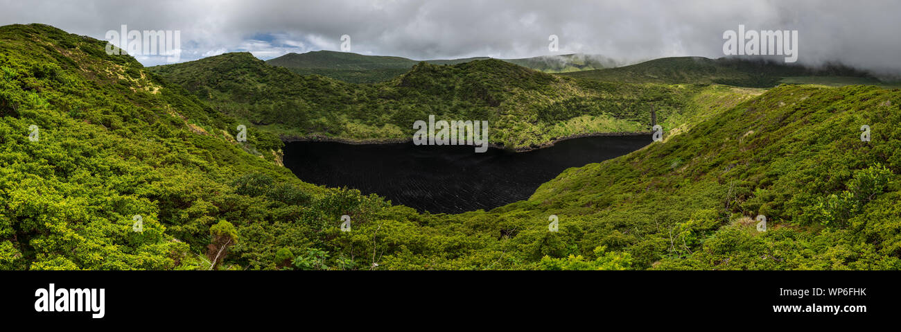 Landscape of Lagoa Comprida caldera crater dark black lake surrounded by green vegetation and with a stormy sky on the Ilha das Flores Island at the A Stock Photo