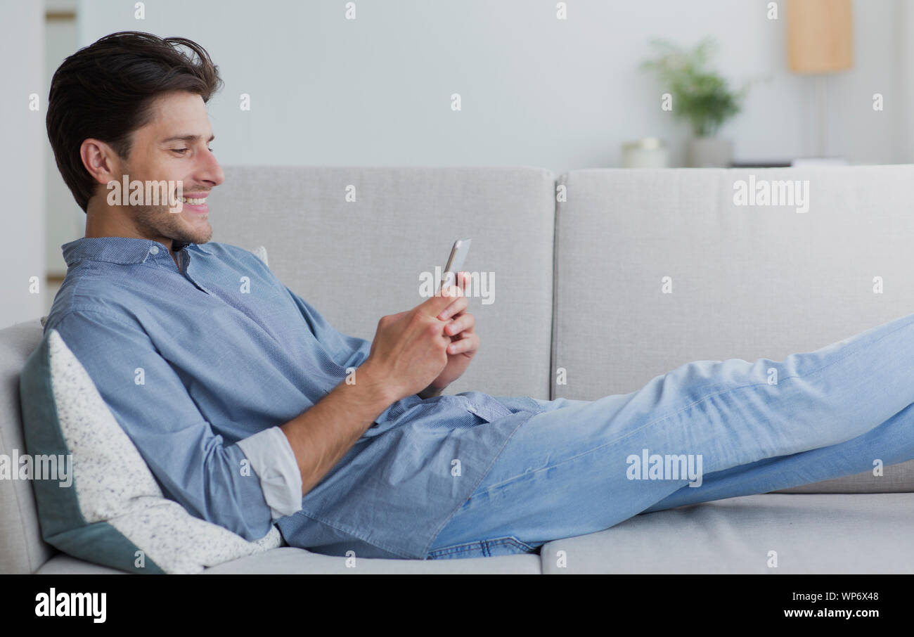 Happy Man Using Smartphone Browsing Internet Lying On Couch Stock Photo