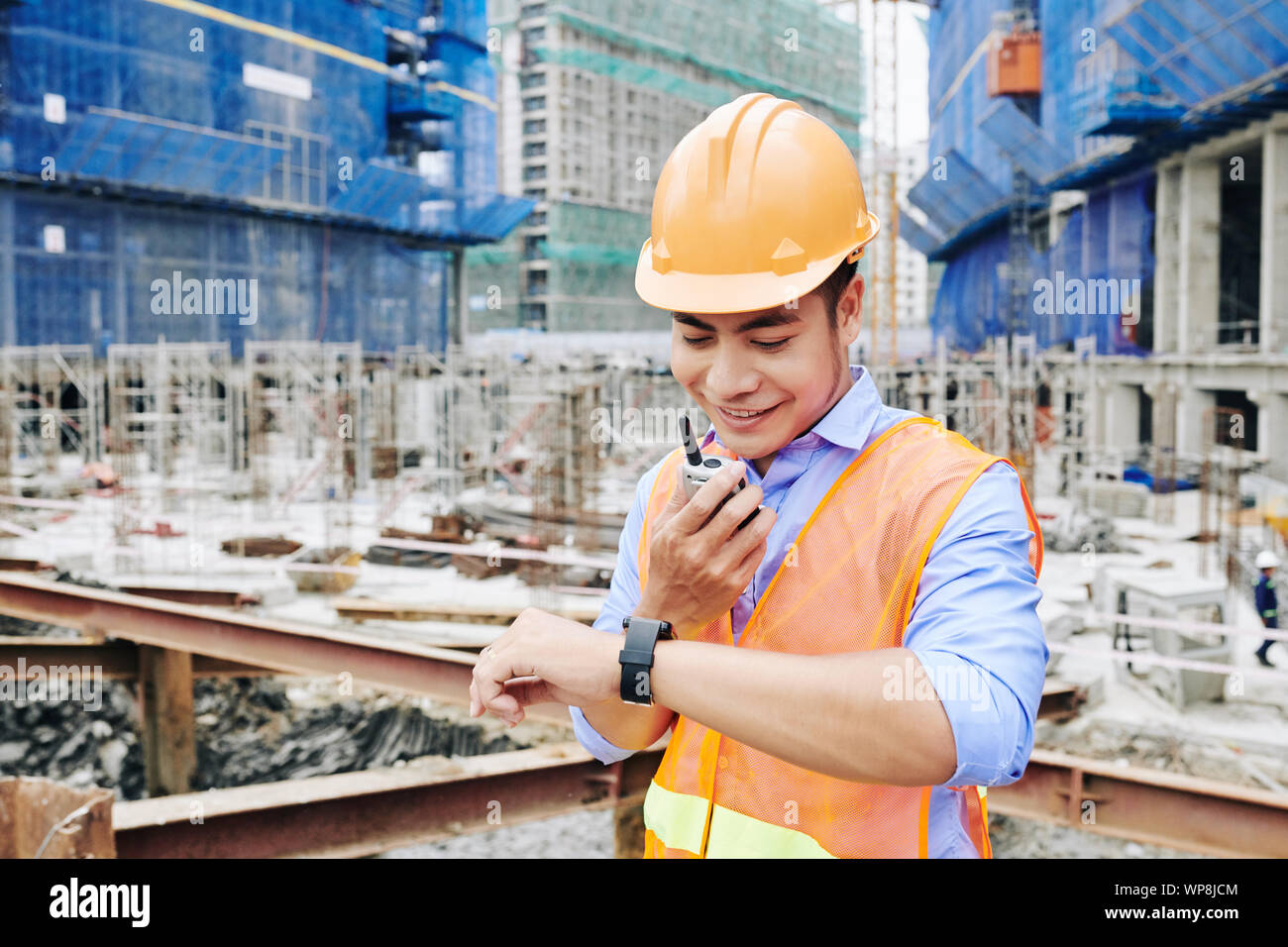 Smiling Asian contractor checking time on his wristwatch when talking to coworkers via walkie-talkie Stock Photo