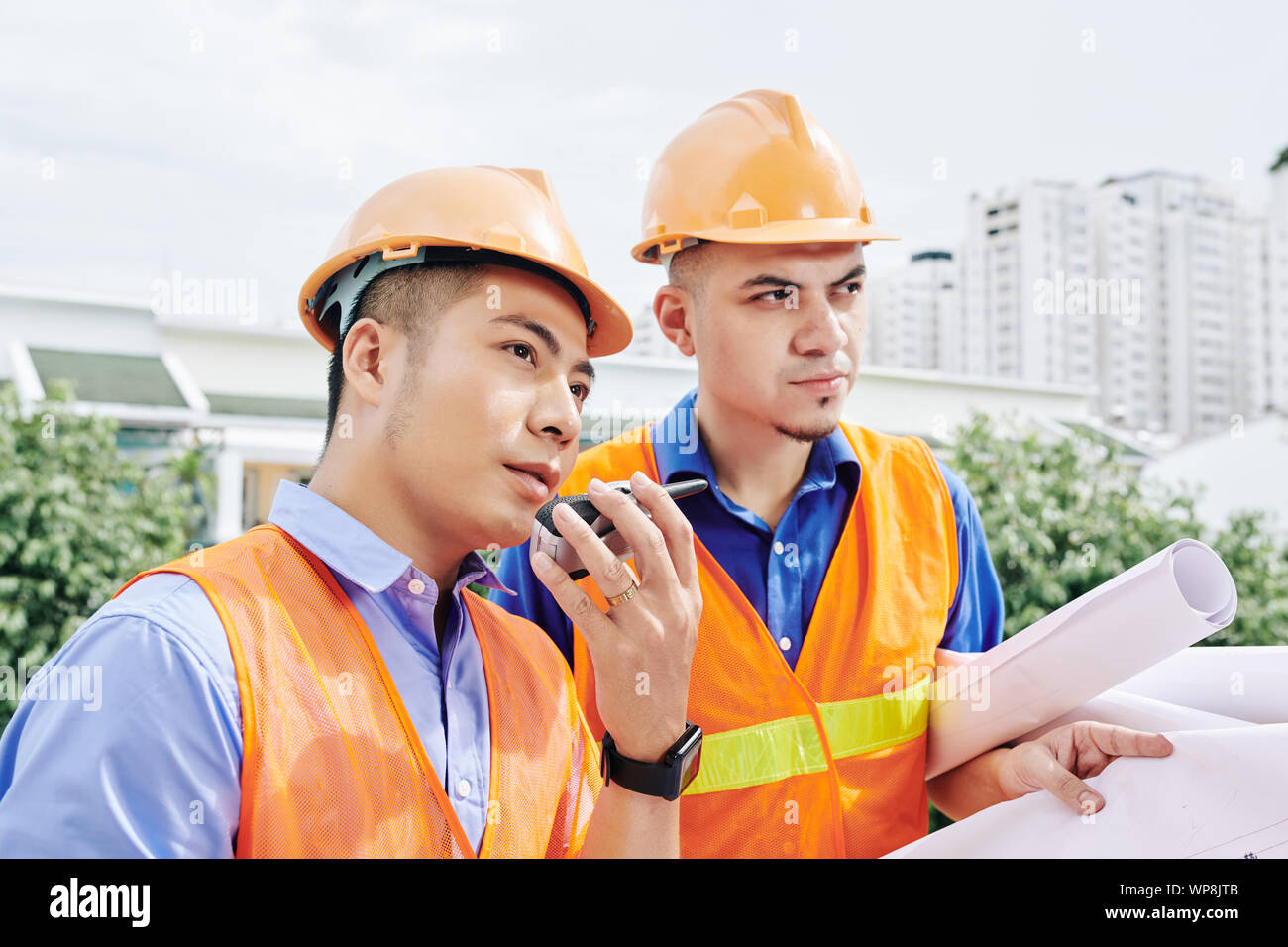 Vietnamese contractor talking on walkie-talkie with workers, his colleague standing nearby with blueprint in hands Stock Photo