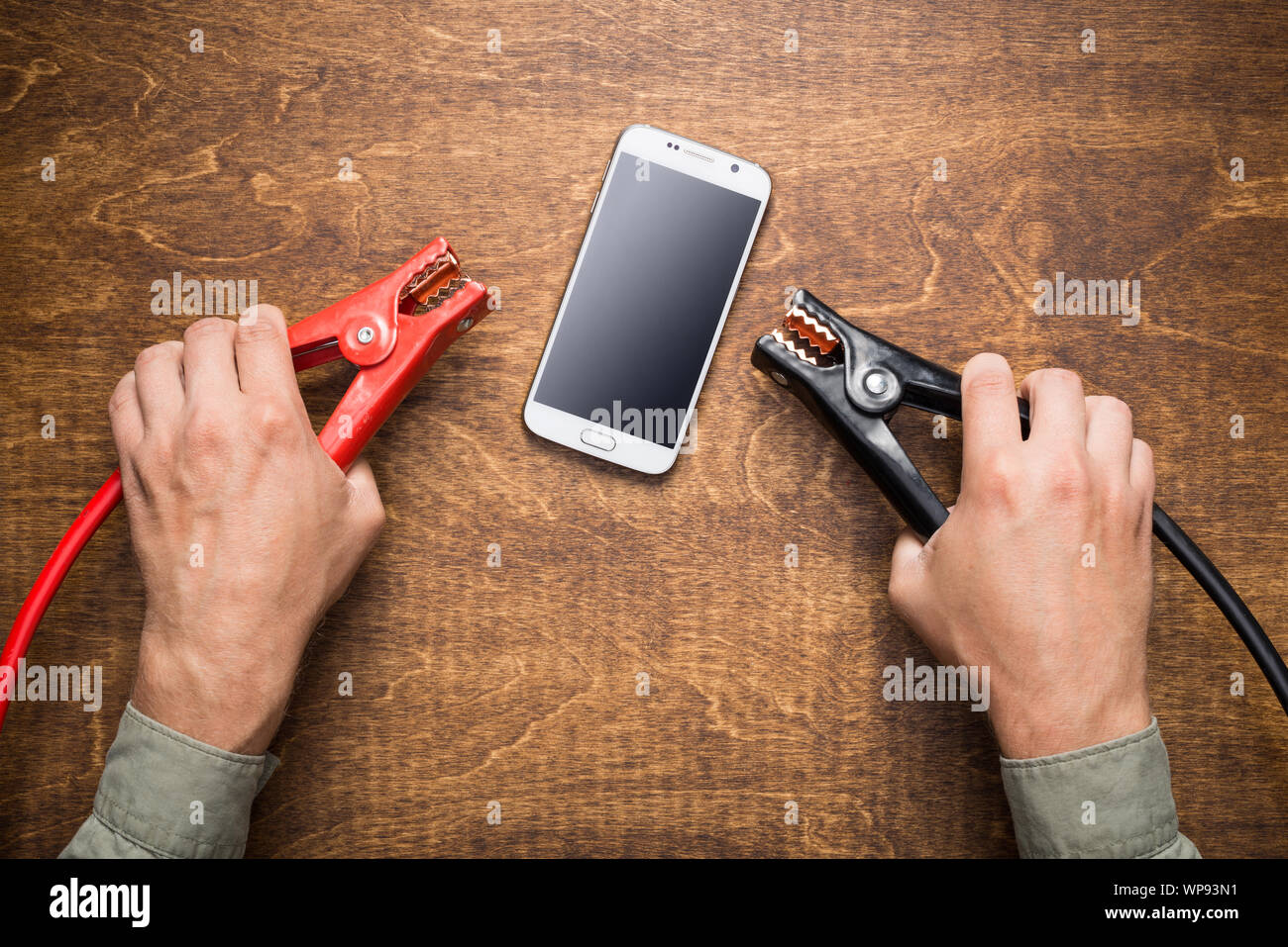 Hands holding charching clamps near smartphone with dead battery. Phone charging, energy efficiency concept. Stock Photo
