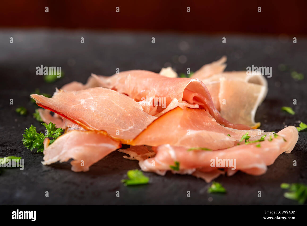 Thin sliced smoked pork ham on a dark slate with herbs Stock Photo