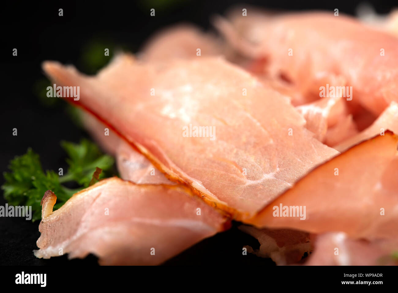 Thin sliced smoked pork ham on a dark slate with herbs - close up view Stock Photo