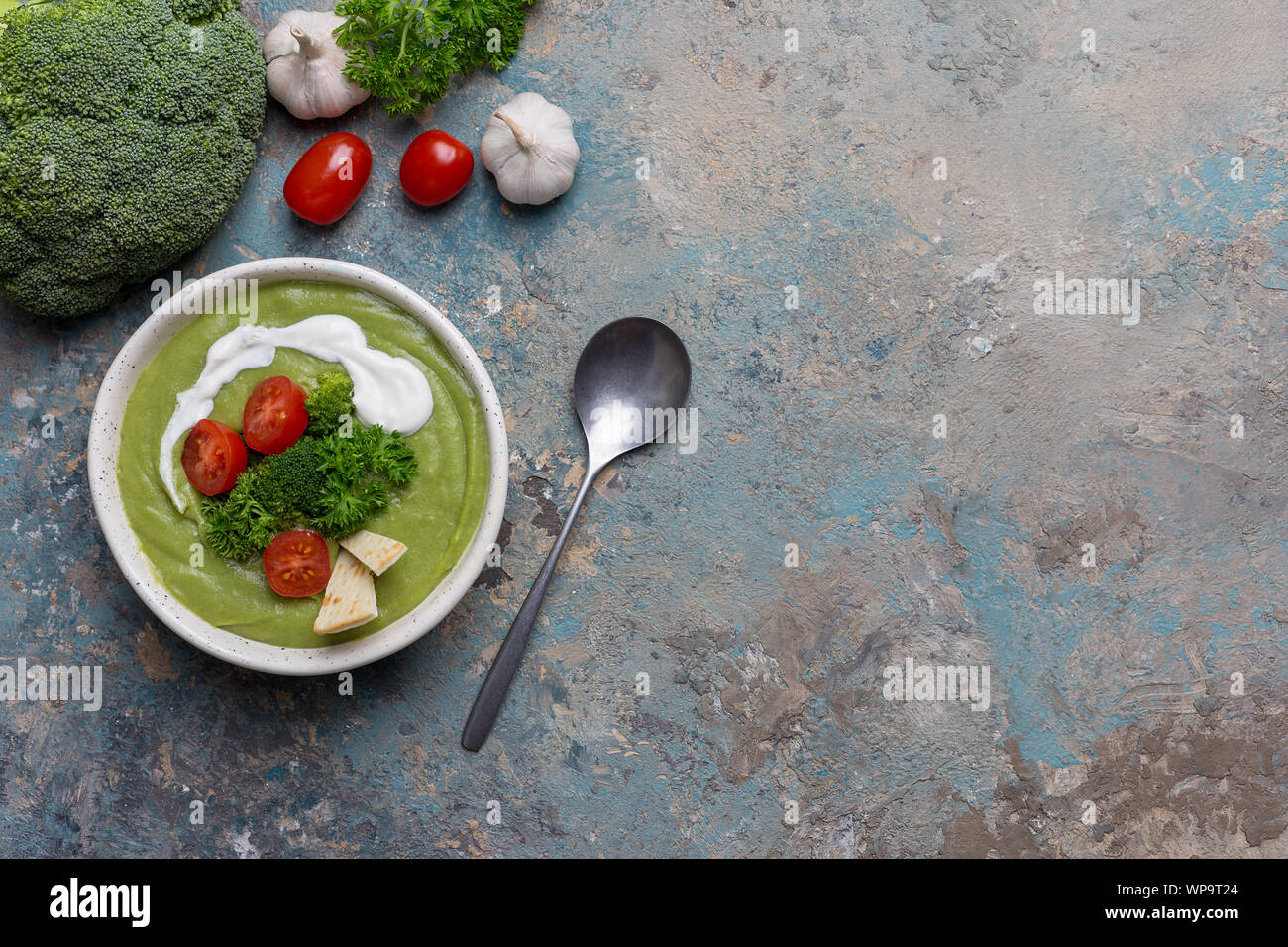 Cream soup with cream and broccoli and raw vegetables as tomatoes, garlic and parsley on blue concrete background. Concept of healthy food. Top view, Stock Photo