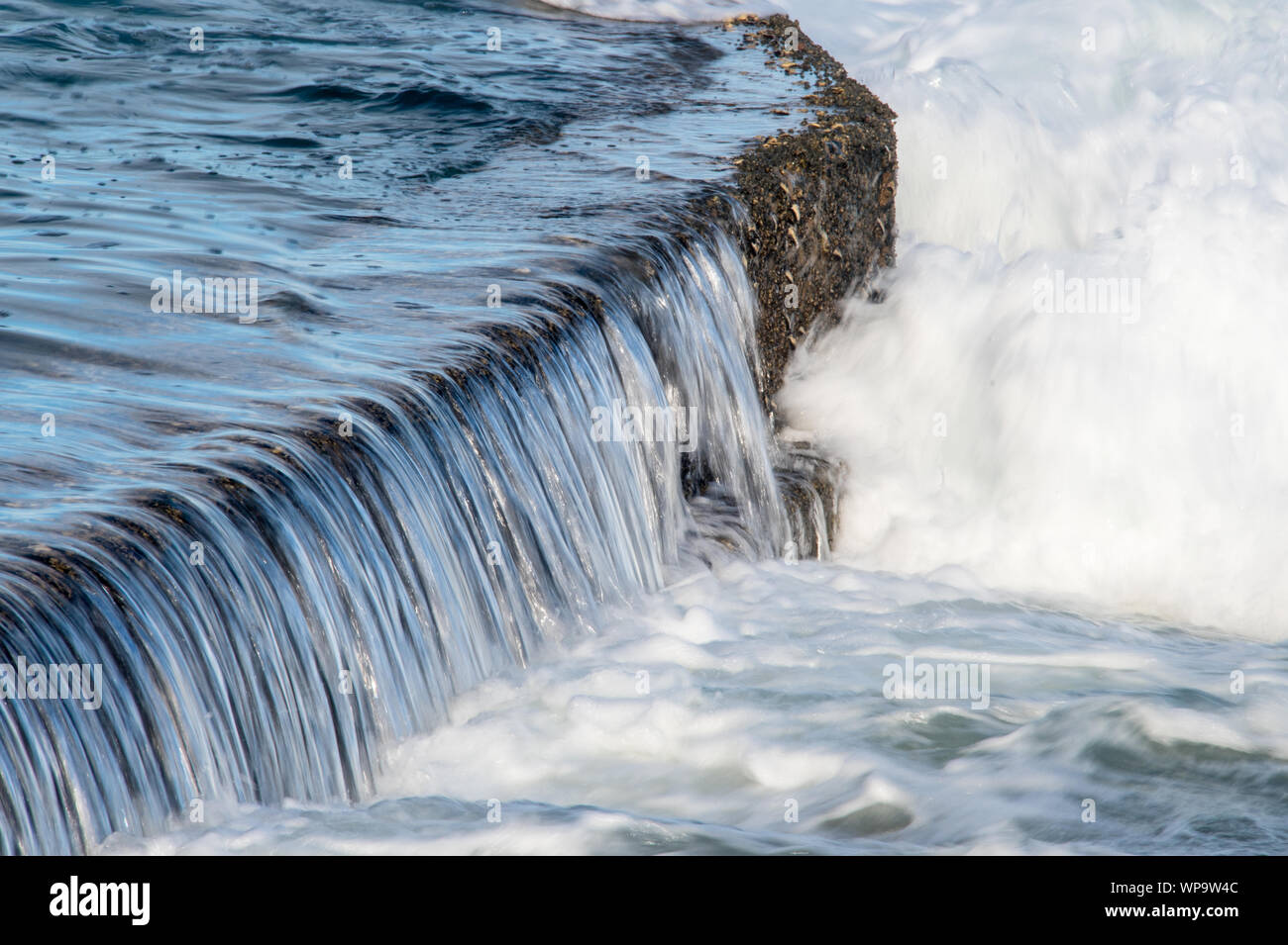 Strong afternoon, incoming tide with waves crashing on a tidal pool wall and creating a powerful overflow foamy surf as backwash crashing Stock Photo