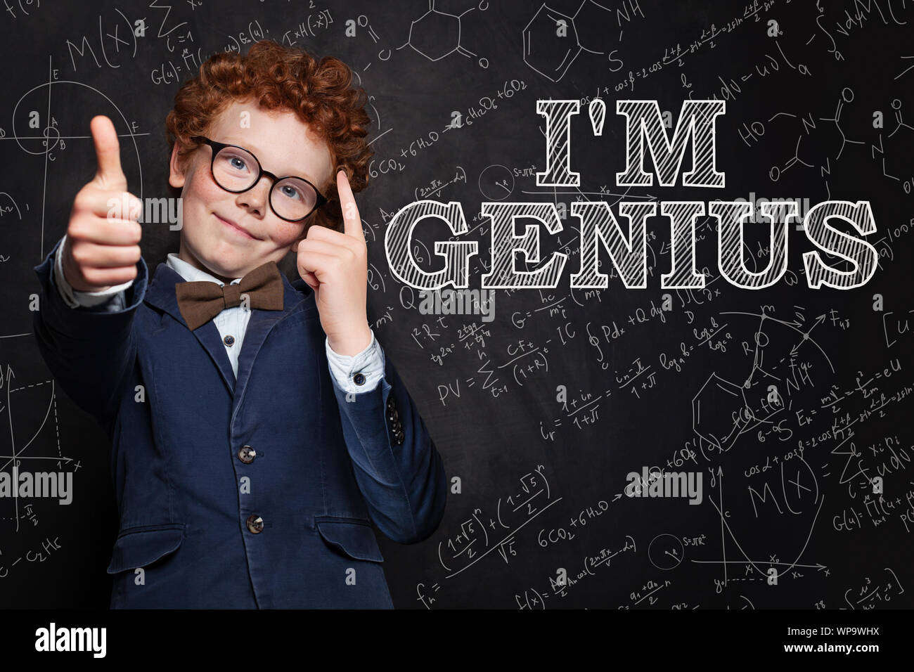 Smiling kid boy with red hair thinking, pointing at his head and standing against blackboard with science formulas Stock Photo