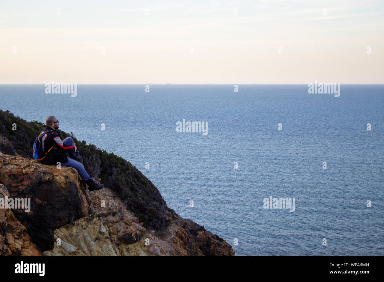 A man is sitting on the end of a high rock and he is looking at the endless sea Stock Photo