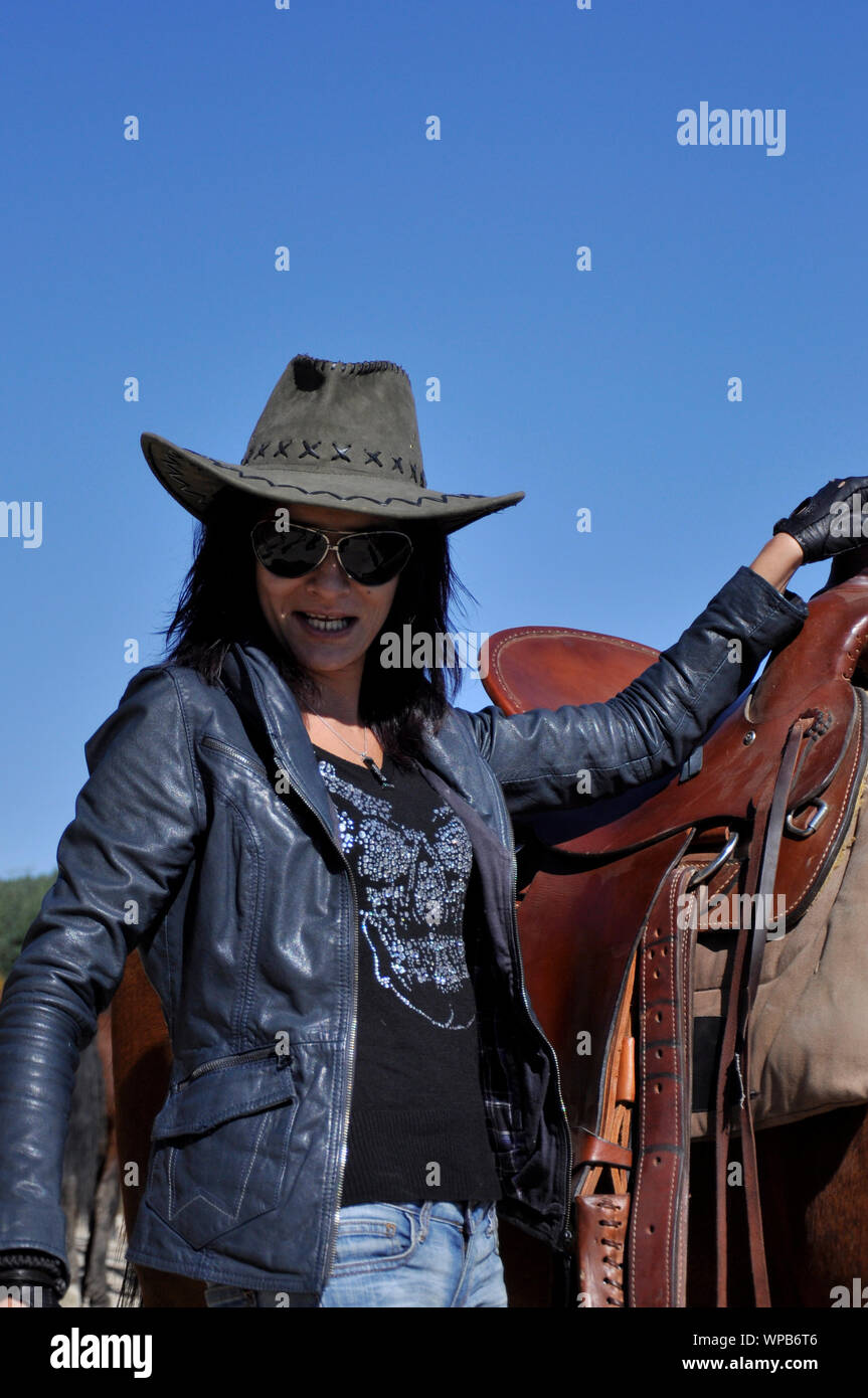 Woman portrait of young Caucasian strong character cowgirl with sunglasses and leather jacket and cowboy hat posing on a ranch by a saddled horse. Stock Photo