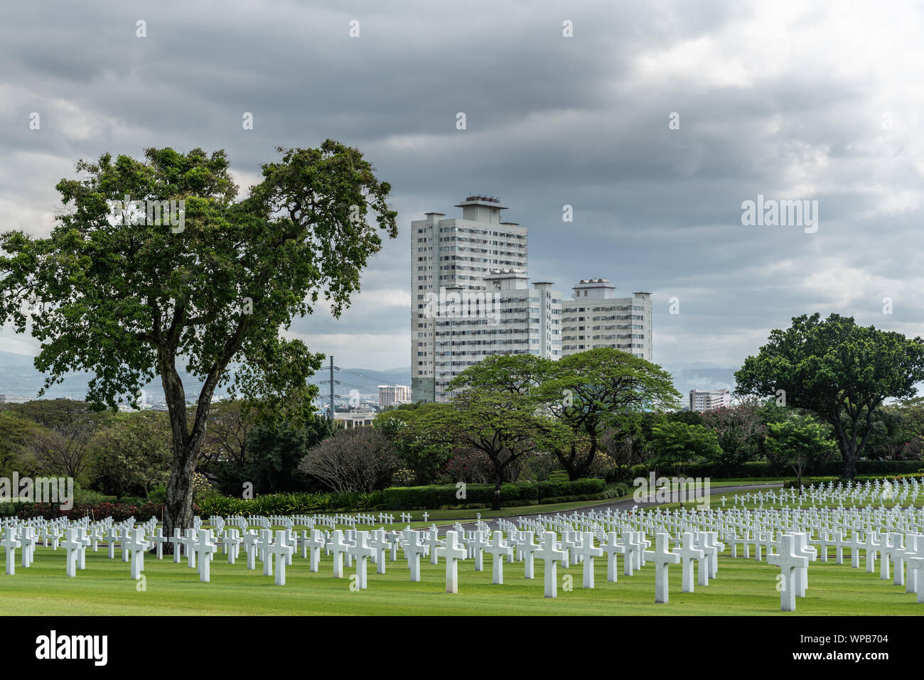 Manila, Philippines - March 5, 2019: American Cemetery and Memorial park. Green lawn, white crosses and heavy cloudscape. White skyscraper on horizon. Stock Photo