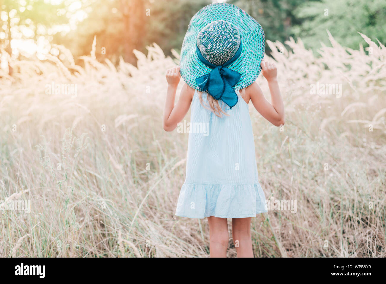 Back view of a cute little girl in a big blue straw hat in the field on ...