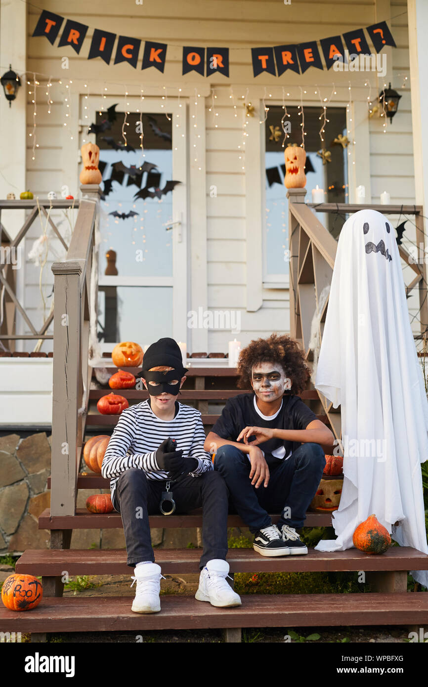 Portrait of two teenage boys wearing Halloween costumes sitting on porch of decorated house and looking at camera, copy space Stock Photo