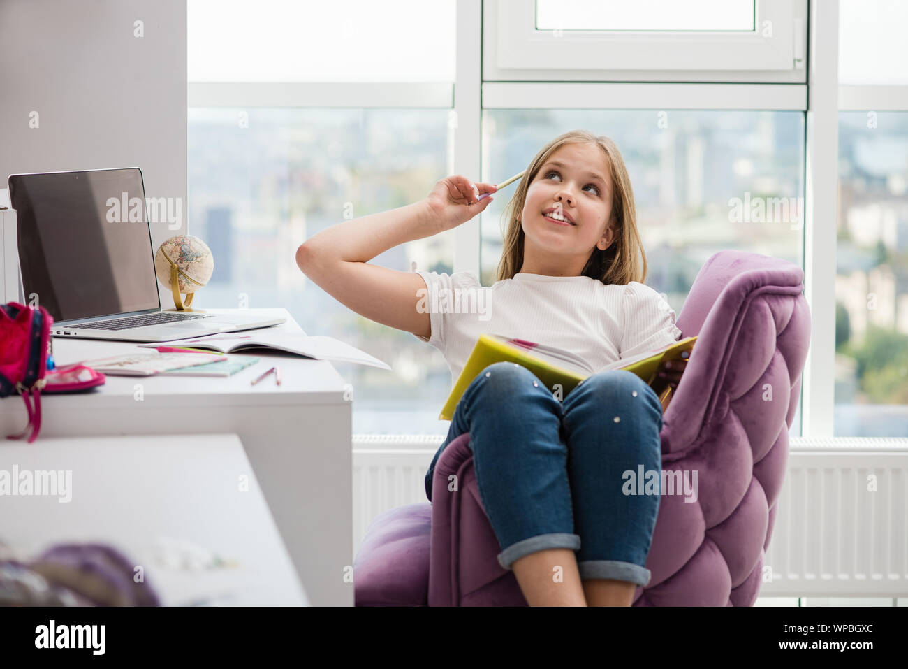 Dreaming pretty school girl with book and pencil. School supplies and laptop in background. Girl sitting in her room with a large window. Stock Photo