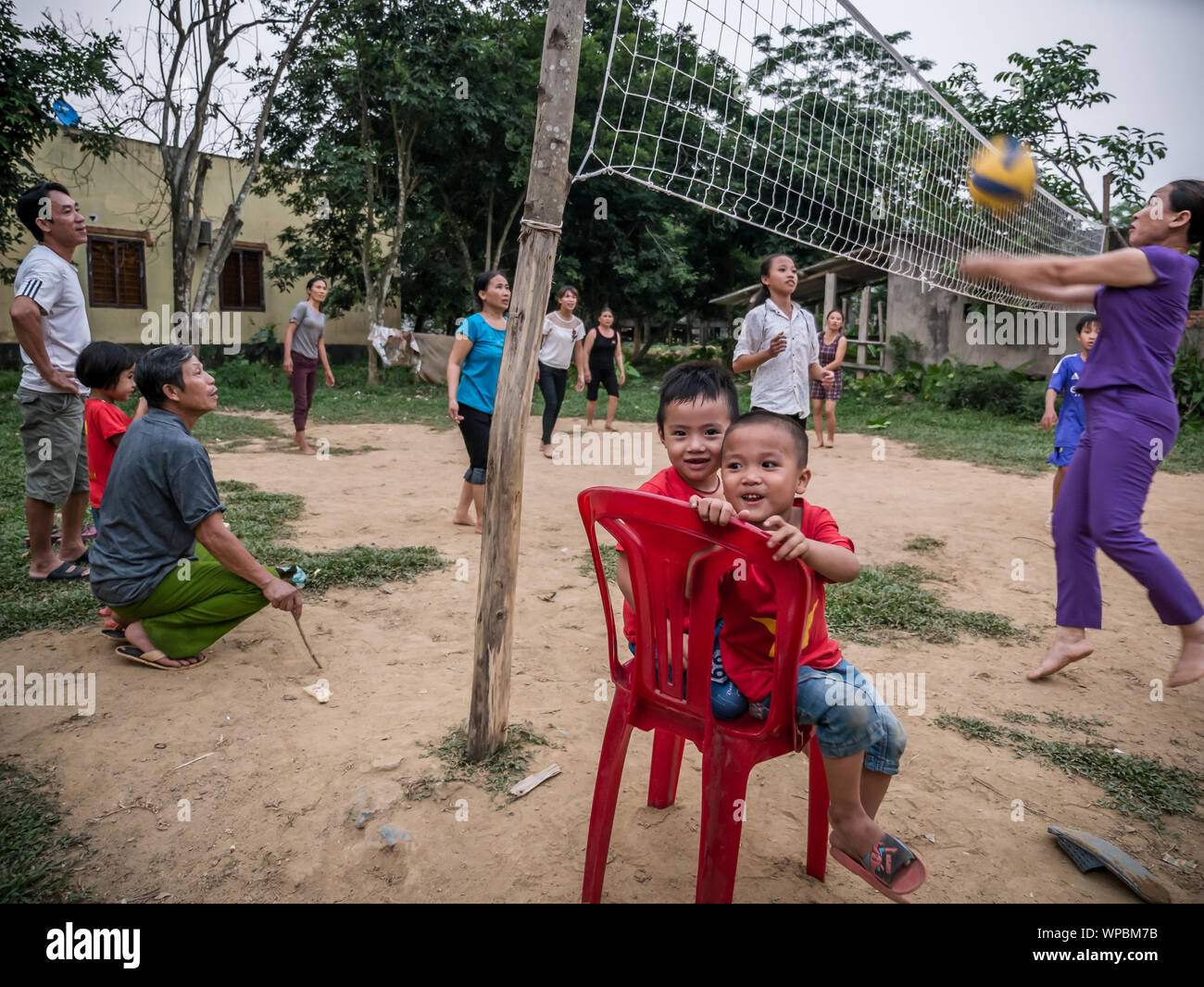 Two young Vietnamese children sitting on red plastic chairs with family and friends playing volleyball in the background, Phong Nha, Vietnam Stock Photo