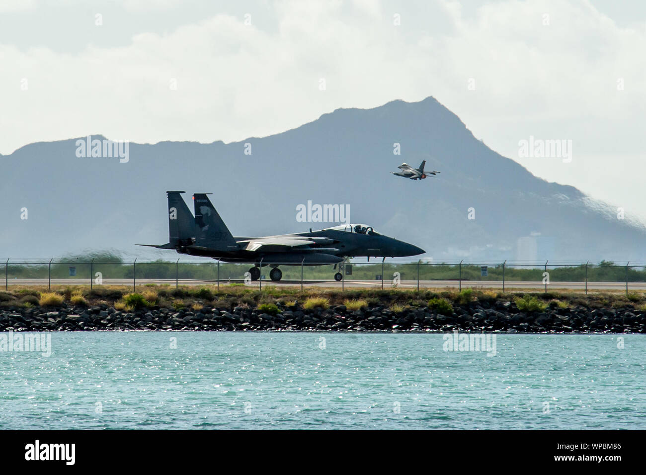An Oregon Air National Guard F-15 Eagle taxis down the Joint Base Pearl Harbor-Hickam flight line Sept. 4, 2019, as an Oklahoma ANG F-16 Fighting Falcon takes off. Fighter and support units from 15 states travel to Hawaii to participate in Sentry Aloha 19-2. The Hawaii Air National Guard exercise is held to provide dissimilar air combat training with fourth and fifth generation Aircraft. (U.S. Air National Guard photo by Senior Airman John Linzmeier) Stock Photo