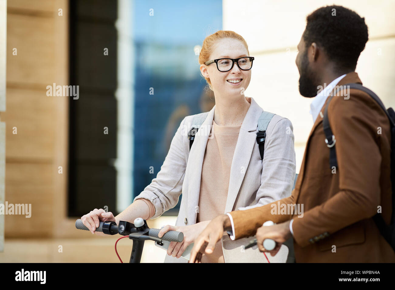 Waist up portrait of modern young couple riding electric scooters in city street, focus on red haired woman smiling cheerfully, copy space Stock Photo