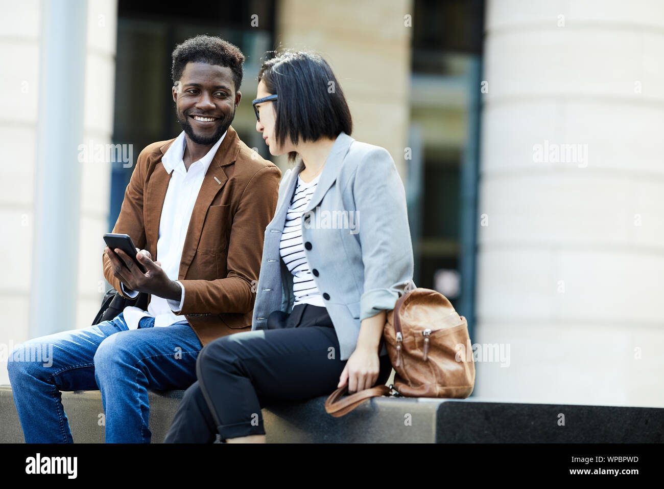 Portrait of two young business people relaxing outdoors during break, African man and Asian woman chatting happily, copy space Stock Photo