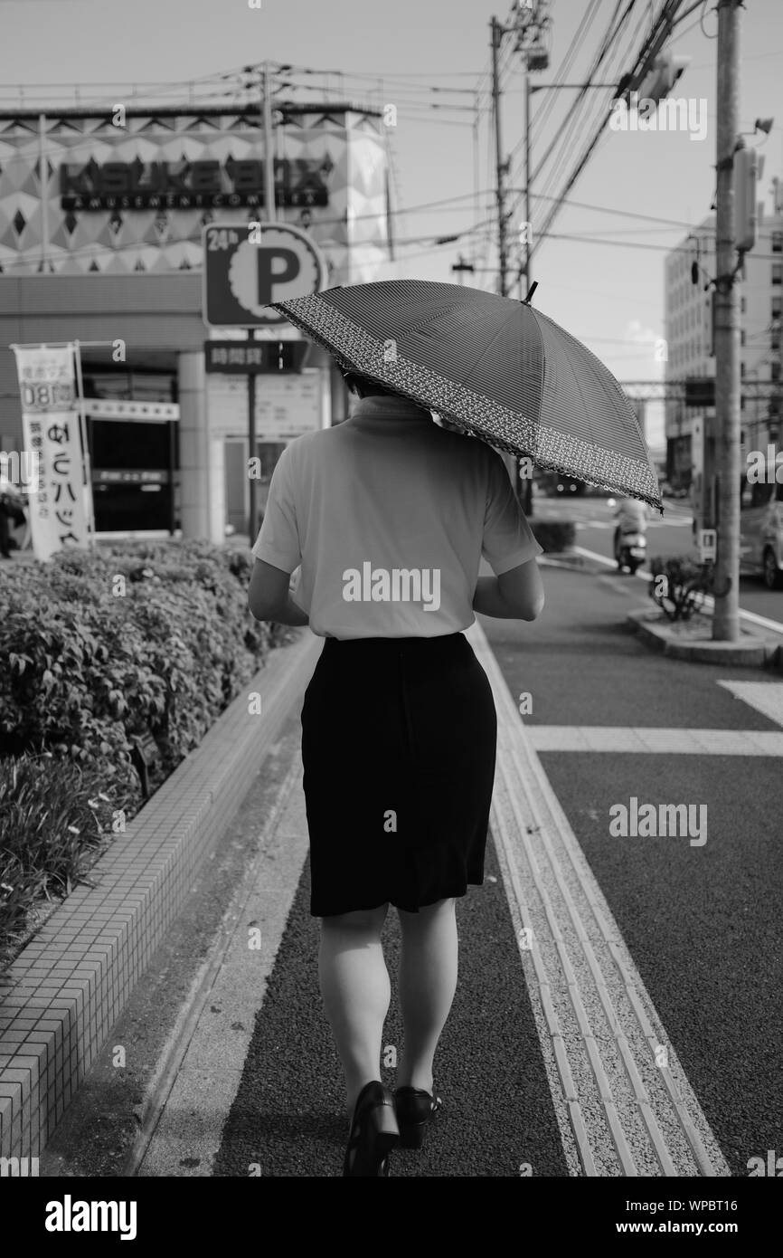 MATSUYAMA, JAPAN - Jul 30, 2019: A grayscale vertical selective shot of a female wearing black skirt with a shirt holding an umbrella walking on a sid Stock Photo