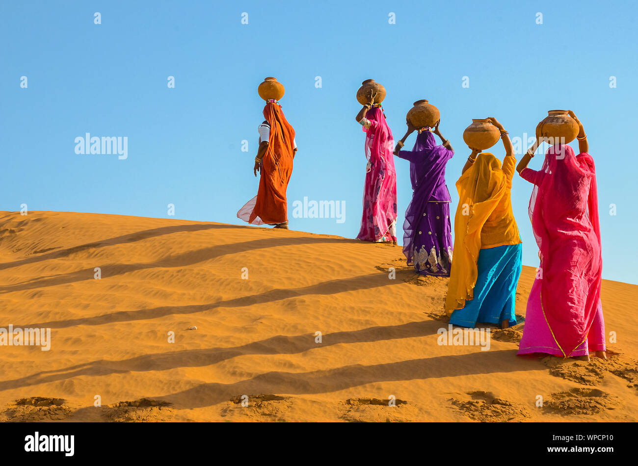 Jaisalmer, rajasthan, india - april 18th, 2018: women carrying heavy jugs of water on their head and walking on a yellow sand dune in the hot summer d Stock Photo