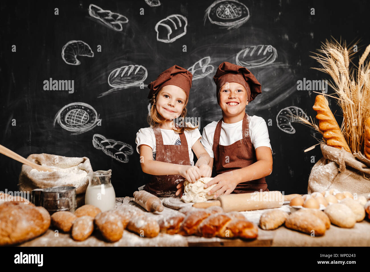 Adorable girl with brother in aprons on table with bread loaves making fresh dough and having fun Stock Photo