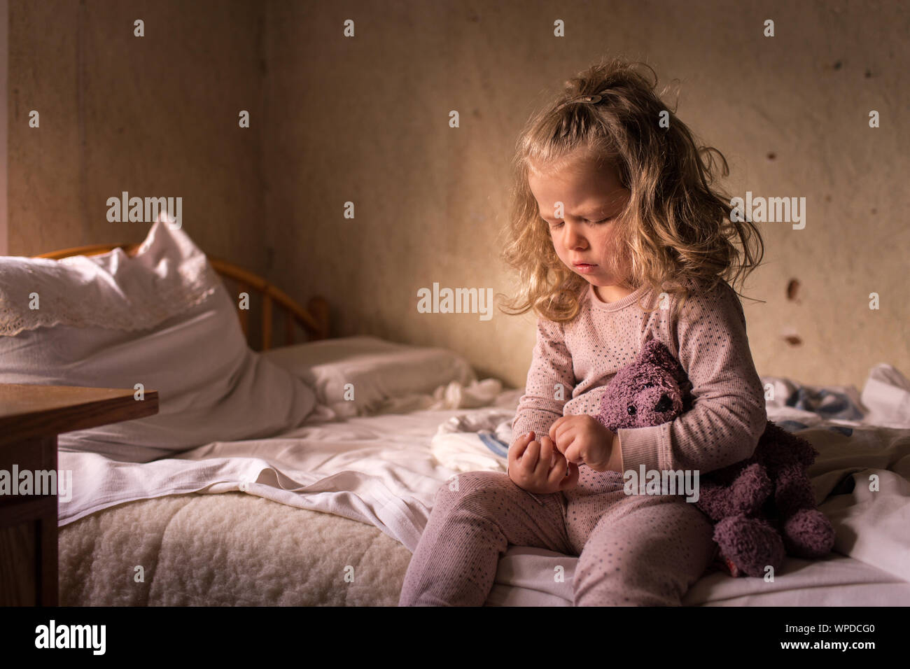 Little kid, sitting on a bed with favorite stuffed toy animal, with serious, worried face expression Stock Photo