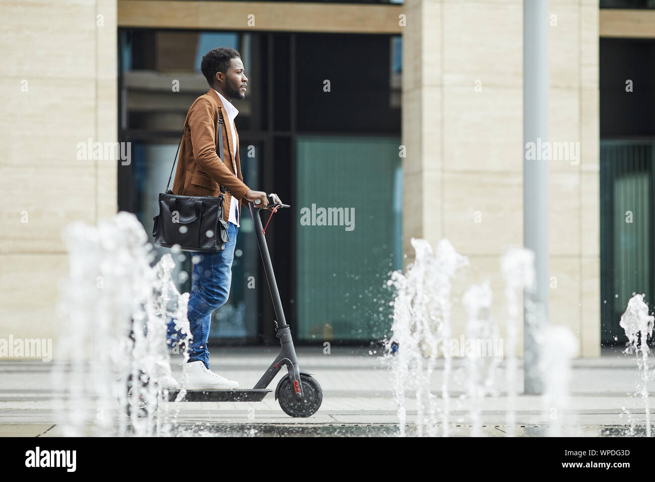 Side view full length of modern African-American man riding electric scooter through fountain while commuting to work in city, copy space Stock Photo