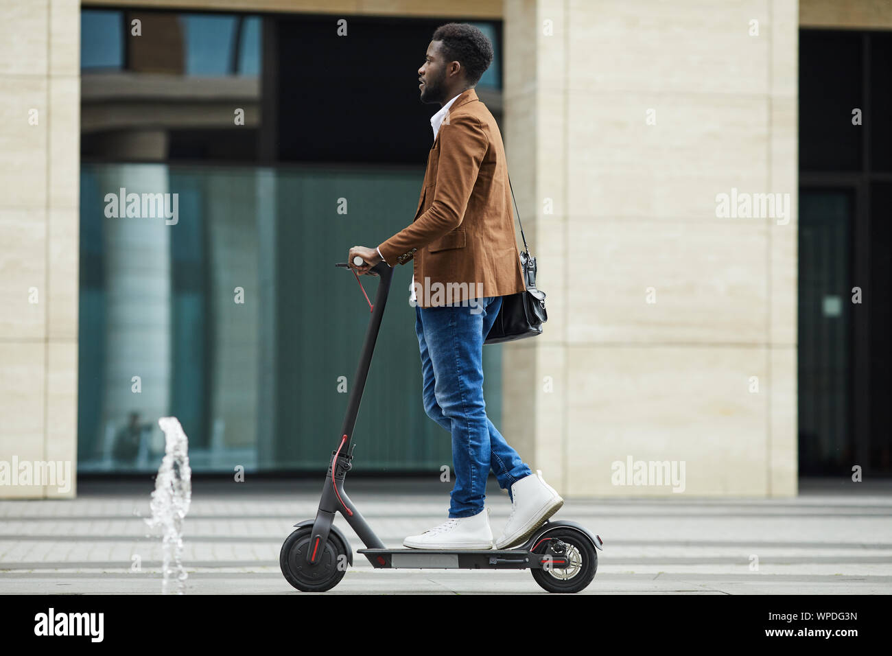 Side view full length of modern African-American businessman riding electric scooter while commuting to work in city, copy space Stock Photo