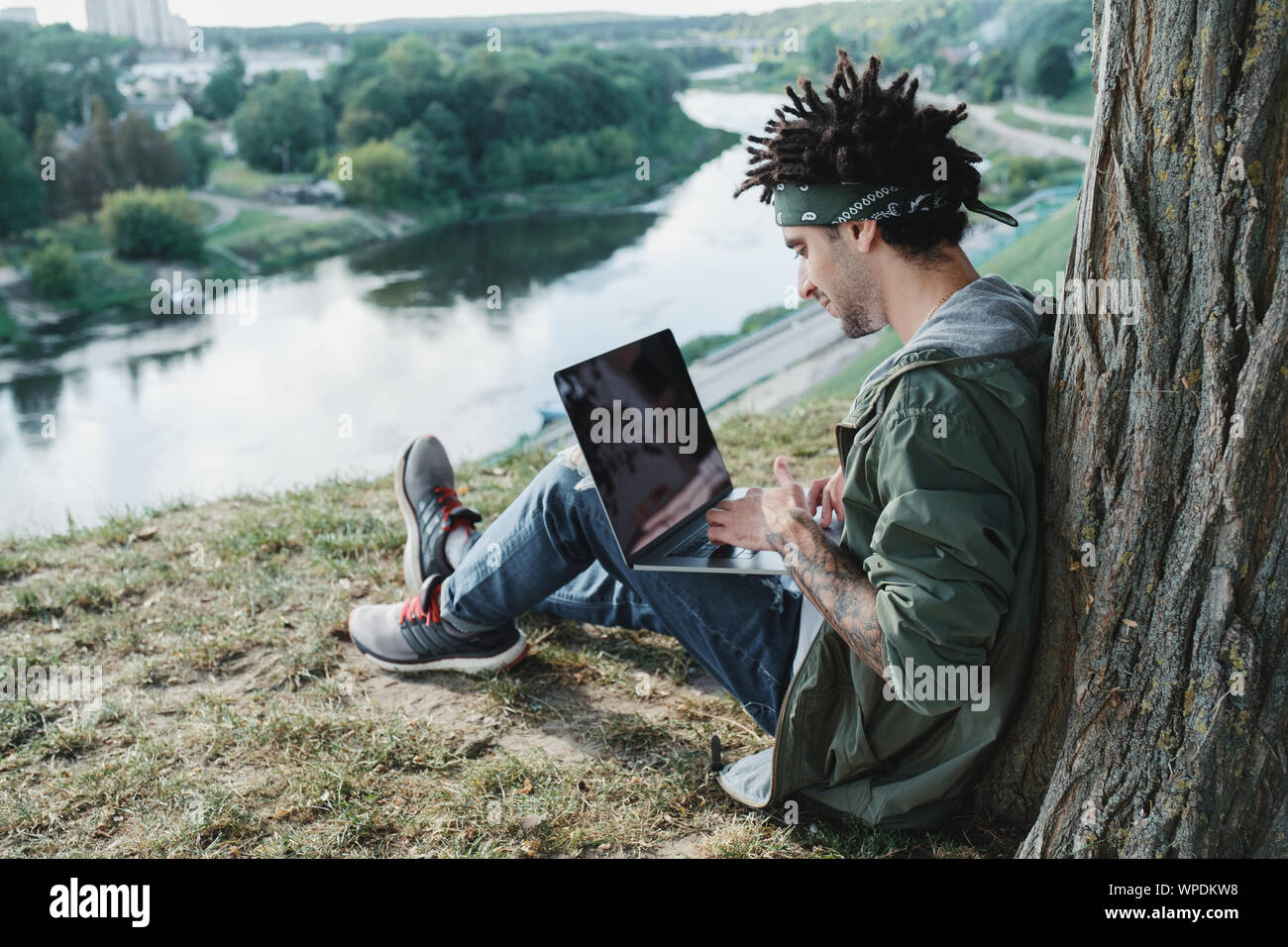 Young millennial student with dreadlocks sitting on grass and using portable laptop computer outdoor in public park. Freelancer using notebook for dis Stock Photo