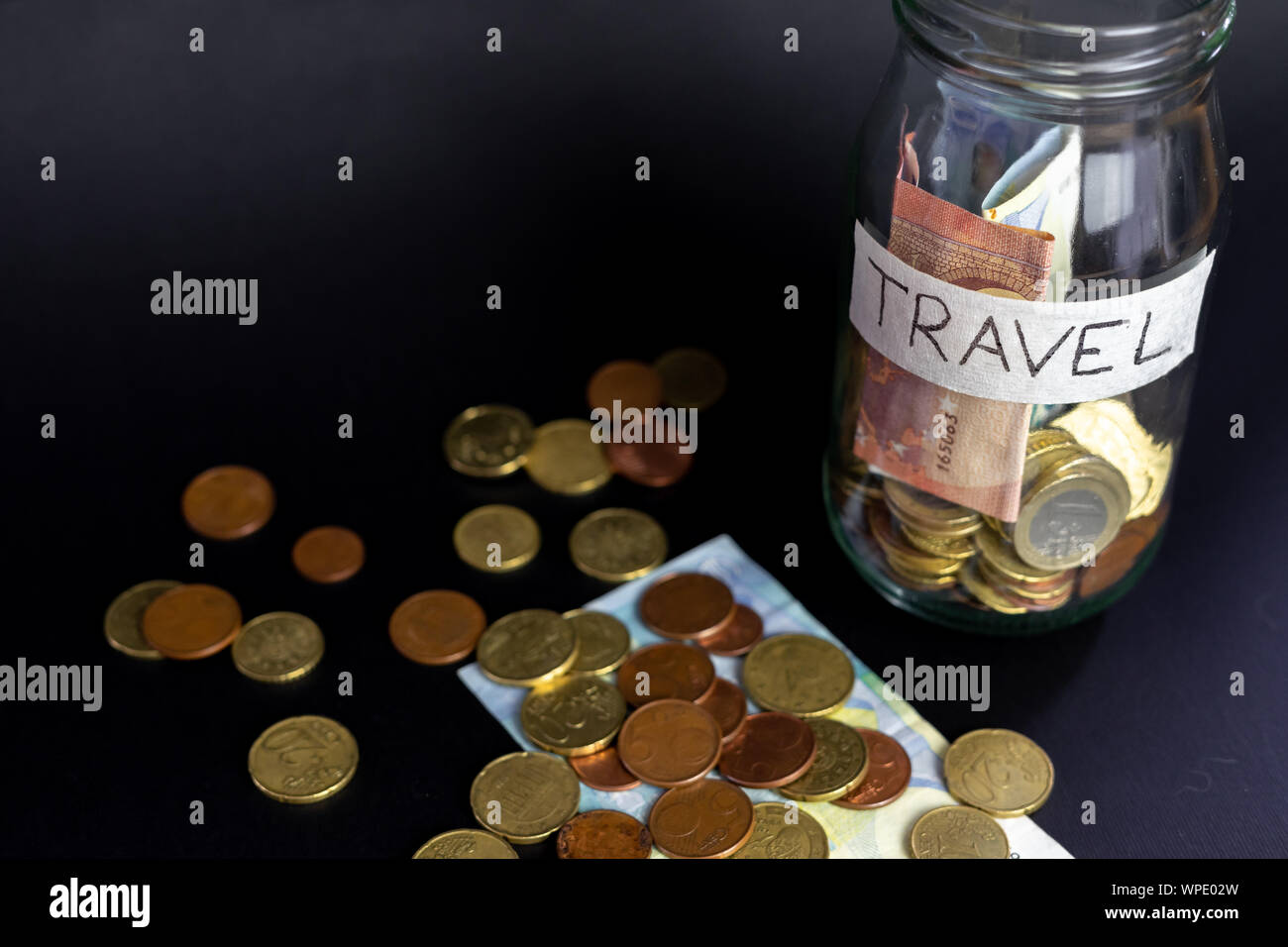 Glass transparent jar labeled handwritten travel. Euro Banknotes and coins inside and outside of the jar. Isolated black background. Stock Photo