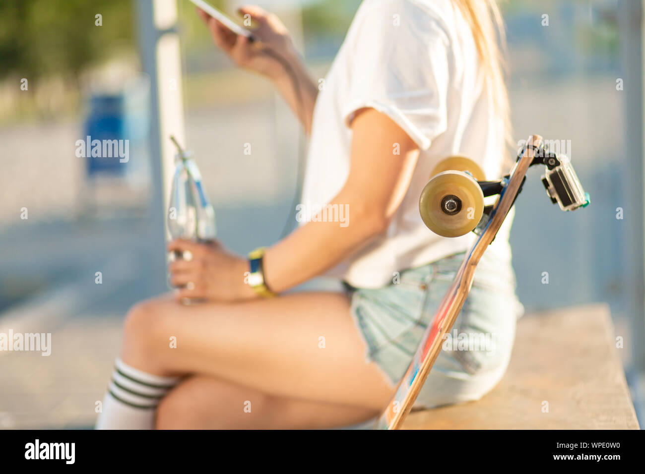 Stylish young girl sitting on bus stop with her longboard, drink soda and wait for bus Stock