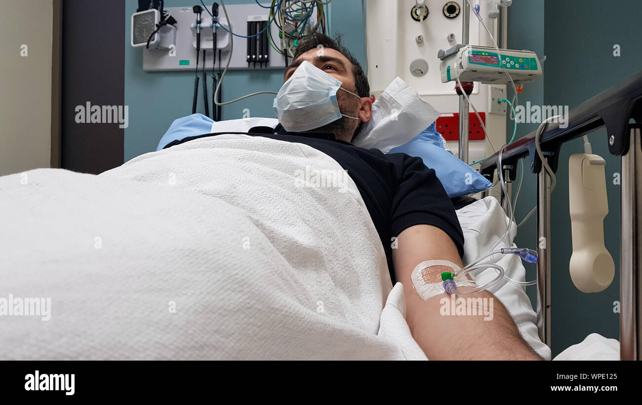 Young man lying in hospital bed. Recovering in modern hospital wards, covered with blanket, face mask and with intravenous needle in his arm Stock Photo